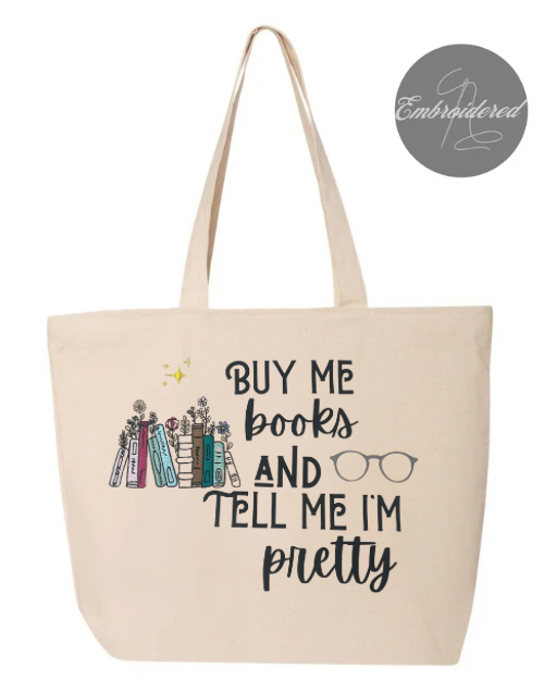 A beige cotton canvas tote bag with the text 'BUY ME BOOKS AND TELL ME I'M PRETTY' printed on the front, along with an illustration of books and glasses.
